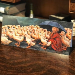 May include: A young girl in a red dress sits under a blue umbrella with a flock of white ducks surrounding her. The ducks are all facing the girl, and she is looking at them with a smile on her face.