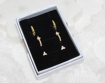 May include: A set of three gold earrings in a black velvet box. The earrings are a pair of small hoops, a pair of small curved earrings with three small stones, and a pair of small stud earrings with three small stones.