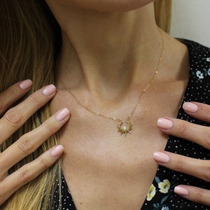 May include: A delicate gold necklace with a sunburst pendant and a central pearl. The necklace is worn by a person wearing a black dress with a floral pattern. The person has fair skin.