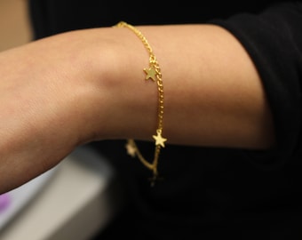 Puede incluir: Un brazalete dorado con dijes de estrella. El delicado brazalete de cadena presenta múltiples colgantes de estrella pequeños. El brazalete se usa en la muñeca de una persona, con un fondo negro.