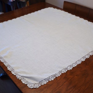 May include: A square, white tablecloth with a delicate lace trim. The tablecloth is made of a soft, woven fabric and is displayed on a dark wooden table. The lace trim adds a touch of elegance.