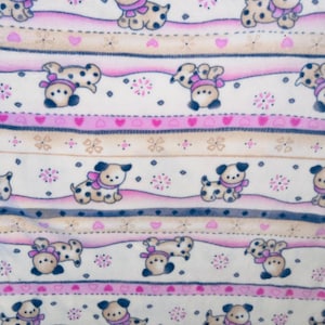May include: A repeating pattern of cartoon dogs with black spots, wearing pink scarves and hats, on a cream background. The design includes pink and tan stripes with hearts and floral accents.