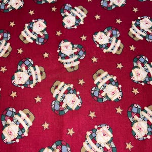 May include: Red fabric with a repeating pattern of Santa Claus figures and gold stars. The Santa figures are in a heart shape, with a white beard, hat, and suit trim. The Santa's suit is a patchwork of blue, green, and red.