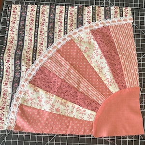 May include: A pink and white fabric quilt with a fan-shaped design. The quilt is made of different patterned fabrics and has a lace trim around the edge.