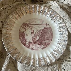 May include: A white ceramic plate with a gold rim and a red and white image of the Suspension Bridge in Royal Gorge, Colorado.
