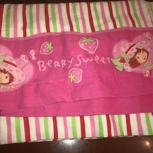 May include: Pink and green striped bed sheet with a pink panel featuring a cartoon strawberry character with the text "Berry Sweet".