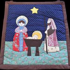 May include: A quilted nativity scene featuring Mary, Joseph, and baby Jesus in a manger. The figures are made of fabric with detailed patterns and colors. The background is a starry night sky with a yellow star.
