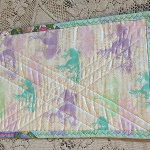May include: A quilted placemat with a light yellow background and a pattern of green, purple, and pink floral designs. The placemat has a light green border with a decorative stitch.