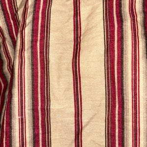 May include: Close-up of a beige fabric with vertical stripes in burgundy, dark gray, and black. The fabric has a textured appearance, with the stripes varying in width and color intensity. The overall effect is a classic, slightly rustic design.