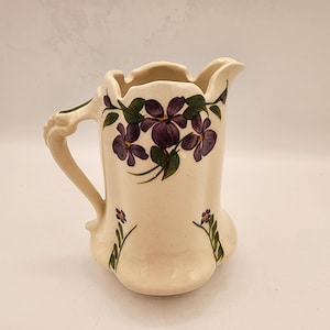 May include: A white ceramic pitcher with a floral design. The pitcher has a handle and a wide mouth. The flowers are purple and green.