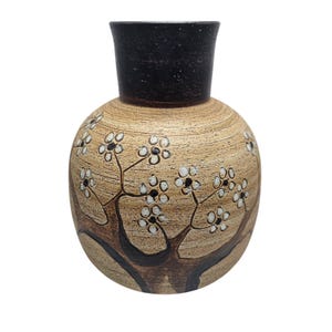 May include: A ceramic vase with a round body and a cylindrical neck. The vase is tan with a dark brown rim and features a hand-painted design of a tree with white flowers and black accents. The vase is a decorative item.