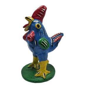 May include: A hand-painted, folk art rooster figurine. The rooster is primarily blue with red, yellow, and green accents. It stands on a green circular base. The rooster's comb and wattle are red, and its beak is yellow.