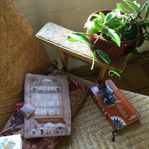 Peut inclure: Un gros plan d'un fauteuil en osier avec un coussin et une broderie représentant un bâtiment avec le texte "Vacanze Romane". Un livre à couverture rouge et une plante dans un pot rouge sont également visibles.