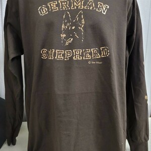 May include: A brown long-sleeve t-shirt with a tan print of a German Shepherd dog's head and the text "GERMAN SHEPHERD" with a copyright symbol and the text "Sew What!" underneath.