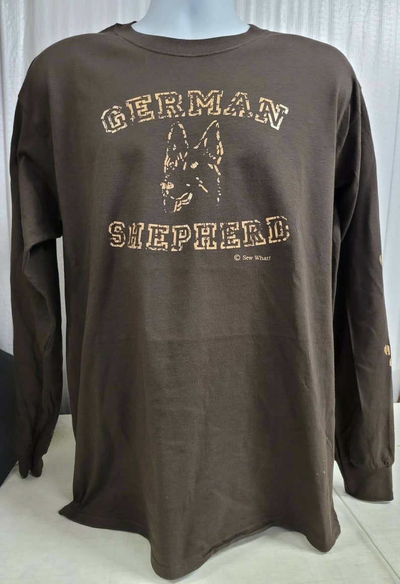May include: A brown long-sleeve t-shirt with a tan print of a German Shepherd dog's head and the text "GERMAN SHEPHERD" with a copyright symbol and the text "Sew What!" underneath.
