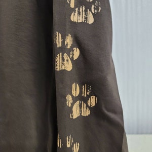 May include: A brown long-sleeved shirt with four paw prints on the sleeve. The paw prints are a light brown color and have a striped pattern.