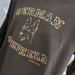 May include: Brown long-sleeve shirt with a tan graphic of a German Shepherd dog and the text "GERMAN SHEPHERD" in a vintage collegiate font. The text "Sew What!" is printed below the graphic.