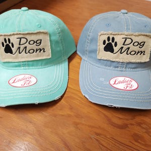 May include: Two denim baseball caps with distressed edges. Both caps feature a beige patch with a black paw print and the text "Dog Mom" in black. The caps are in light blue and turquoise colors. Each cap has a red oval tag that says "Ladies Fit".