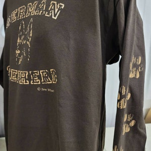 May include: Brown long-sleeve t-shirt with a gold print of a German Shepherd dog and the text "GERMAN SHEPHERD" with the copyright symbol and "Sew What!" underneath. The right sleeve has a gold print of paw prints.