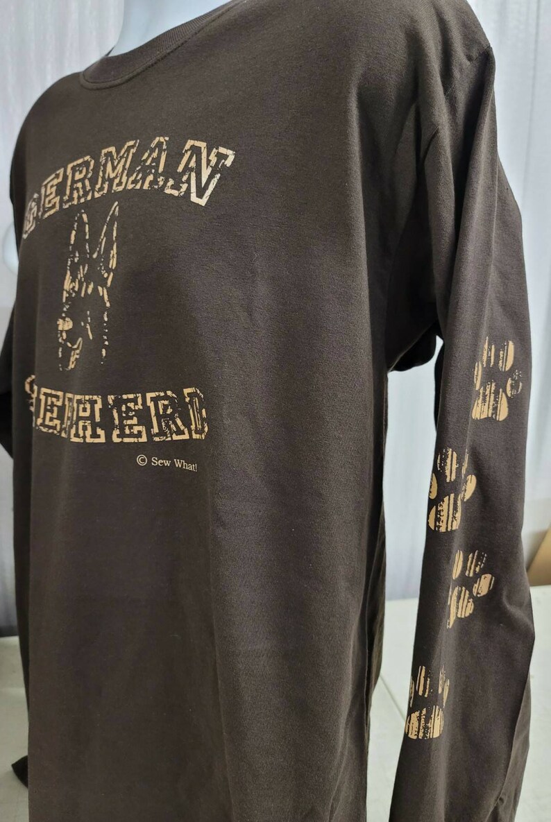 May include: Brown long-sleeve t-shirt with a gold print of a German Shepherd dog and the text "GERMAN SHEPHERD" with the copyright symbol and "Sew What!" underneath. The right sleeve has a gold print of paw prints.