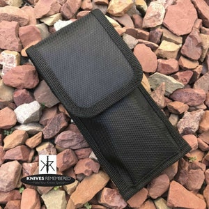 May include: Black nylon sheath for a knife. The sheath has a zipper closure and a belt loop. The sheath is sitting on a bed of small red and brown rocks.