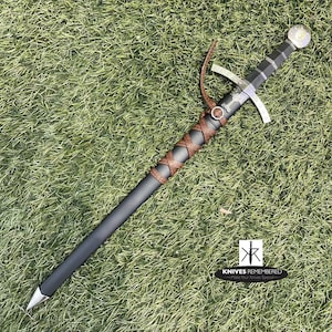 May include: A black and silver sword with a brown leather wrapped handle. The sword has a cross guard and a pommel with a decorative design. The sword is lying on a green grassy surface.