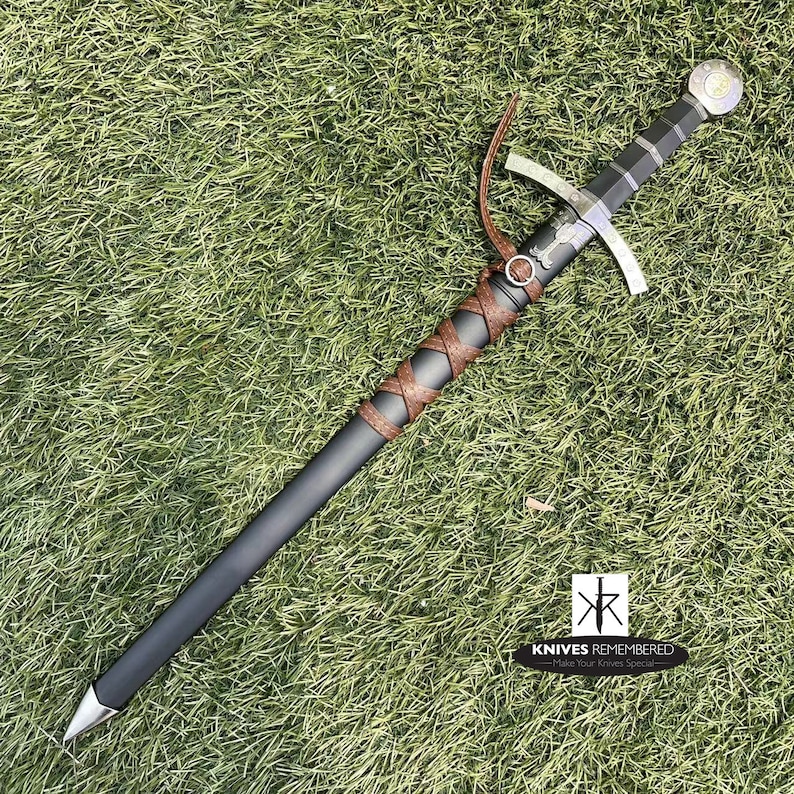 May include: A black and silver sword with a brown leather wrapped handle. The sword has a cross guard and a pommel with a decorative design. The sword is lying on a green grassy surface.