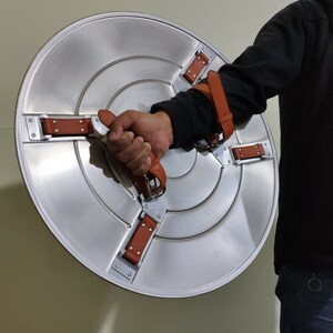 May include: A silver metal shield with brown leather straps. The shield has a raised center and is held by a hand.