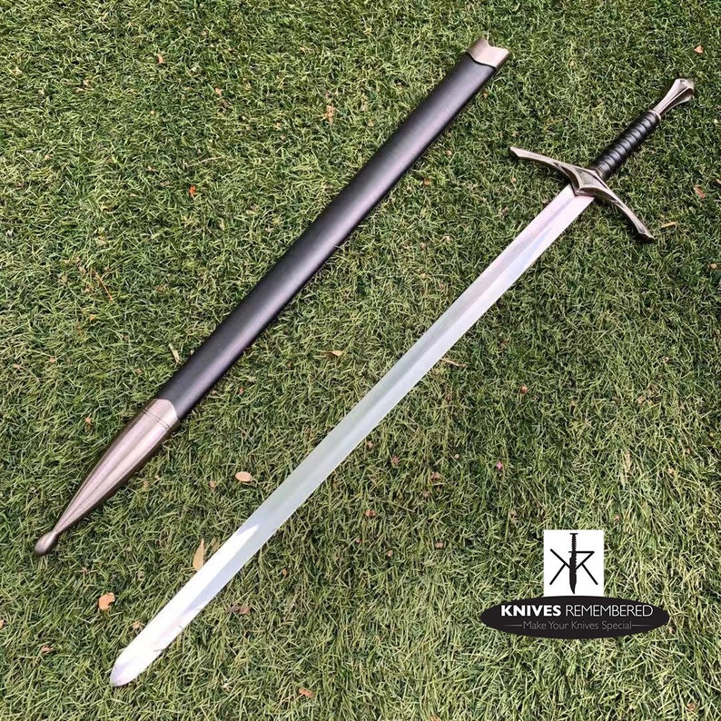 May include: A silver sword with a black handle and a black leather scabbard. The sword is lying on a green grassy surface.
