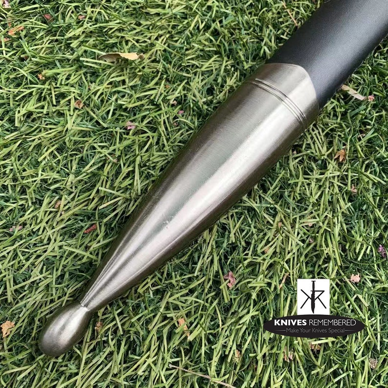 May include: Close-up of the pommel of a sword with a silver finish. The pommel is round and has a smooth surface. The sword is resting on a bed of green grass.