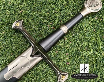 May include: A silver sword with a black leather grip and a gold-coloured pommel. The pommel features a lion design. The sword is lying on a bed of green grass.