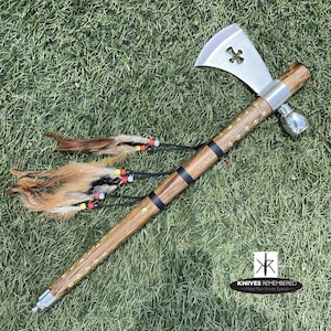 May include: A wooden and metal tomahawk pipe with a brown wooden handle, black accents, and a silver head with a cross-shaped hole. The pipe has a decorative feather and bead tassel.