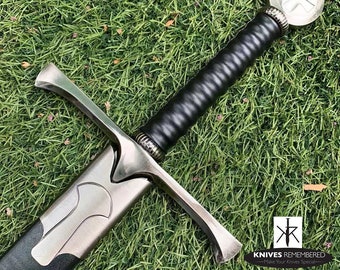 May include: A silver sword with a black leather grip and a cross-shaped pommel. The sword is lying on a bed of green grass.