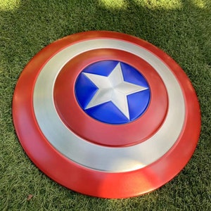 May include: A red, white, and blue Captain America shield with a silver rim. The shield is lying on a green grassy surface.
