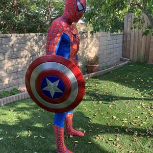 May include: A person dressed as Spiderman is holding a red, white, and blue Captain America shield. The shield is round and has a star in the center. The person is standing on a green lawn with brown leaves scattered around.