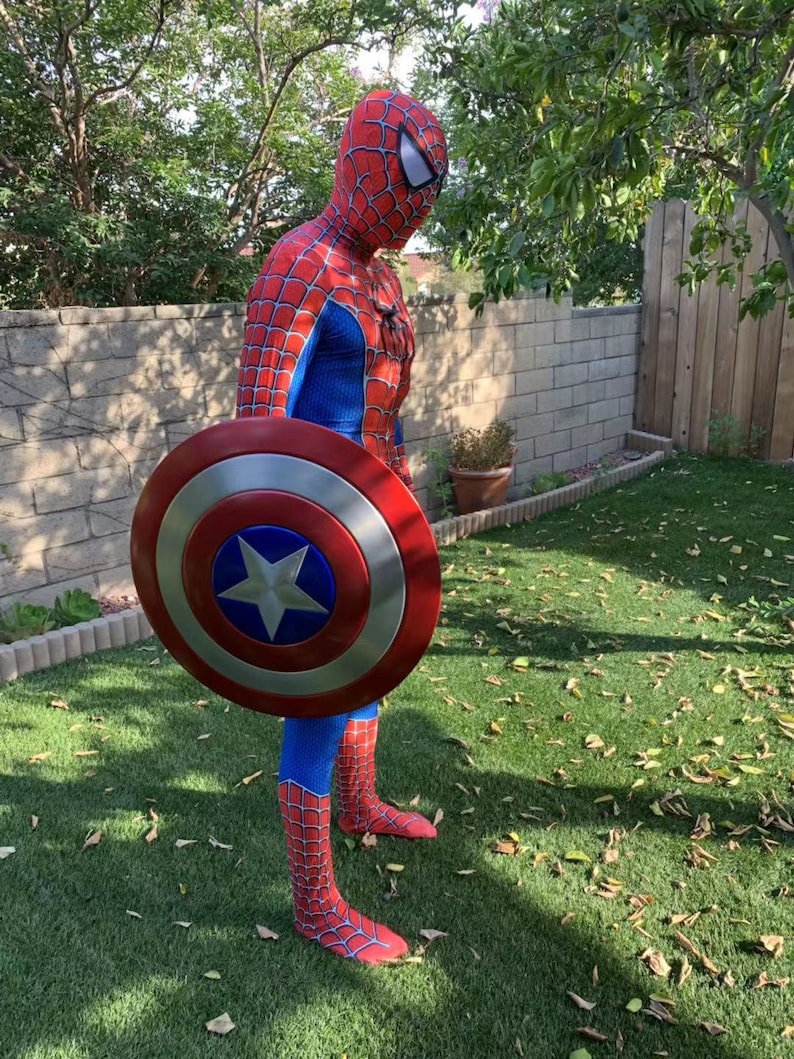 May include: A person dressed as Spiderman is holding a red, white, and blue Captain America shield. The shield is round and has a star in the center. The person is standing on a green lawn with brown leaves scattered around.