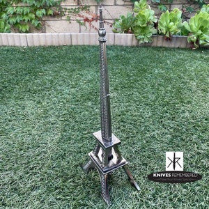 May include: A silver metal Eiffel Tower replica, standing on a green grassy surface. The tower is detailed with intricate designs and has a pointed spire. The text "KNIVES REMEMBERED - Make Your Knives Special -" is visible in the bottom right corner of the image.