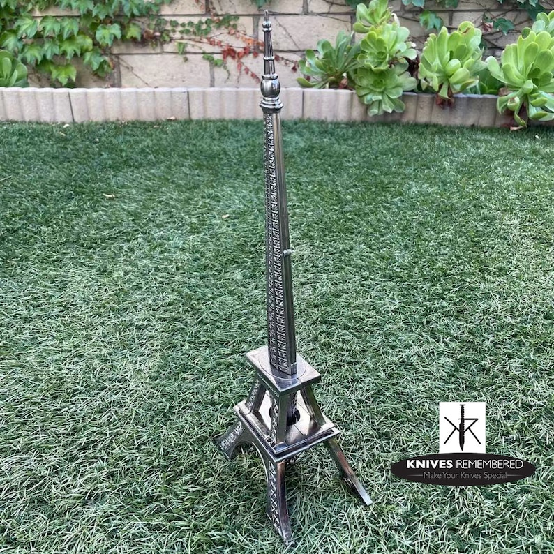 May include: A silver metal Eiffel Tower replica, standing on a green grassy surface. The tower is detailed with intricate designs and has a pointed spire. The text "KNIVES REMEMBERED - Make Your Knives Special -" is visible in the bottom right corner of the image.