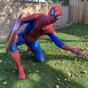 May include: A person dressed as Spiderman in a red and blue costume with a spider web design is kneeling on green grass. The person is holding a red, white, and blue shield in their left hand.