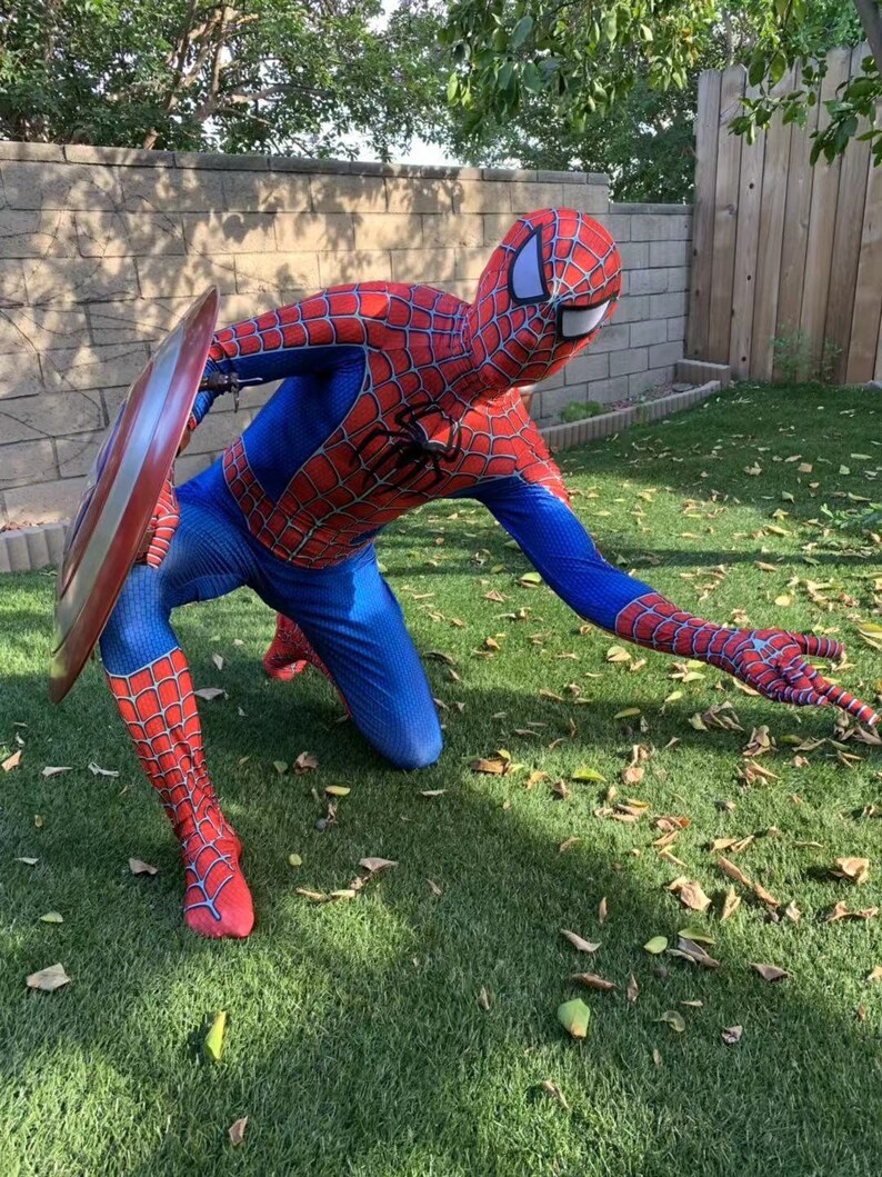 May include: A person dressed as Spiderman in a red and blue costume with a spider web design is kneeling on green grass. The person is holding a red, white, and blue shield in their left hand.