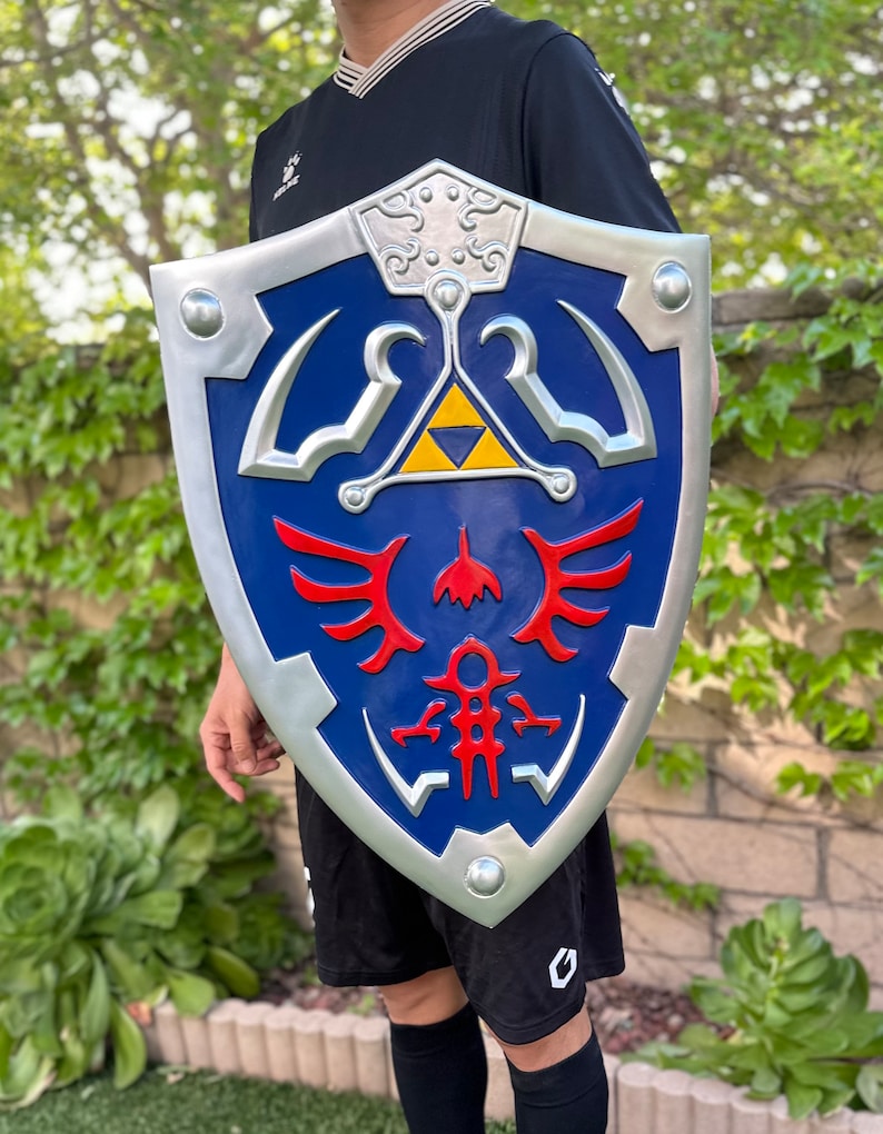 May include: A silver and blue shield with a red and yellow design. The shield has a triangular design with a winged symbol and a stylized figure.