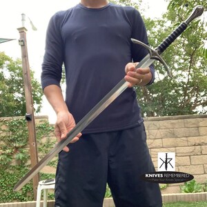 May include: A person holding a silver sword with a black handle. The sword has a cross guard and a pointed tip. The person is wearing a dark blue shirt and black shorts. The background is a blurry image of a wooden fence and green grass. The image also includes the text "KNIVES REMEMBERED - Make Your Knives Special - "