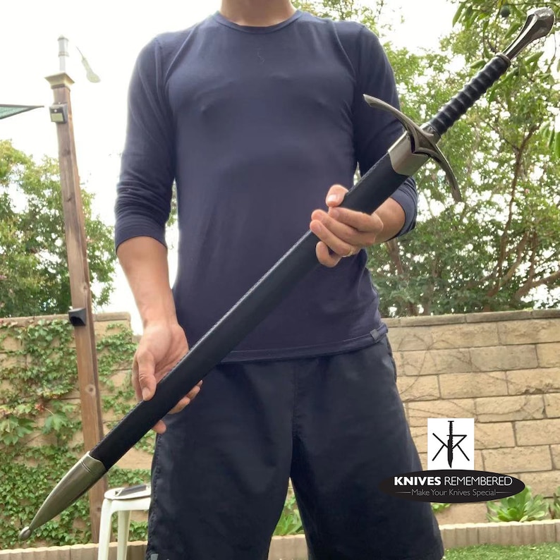 May include: A black leather-wrapped sword with a silver hilt and a black scabbard. The sword is held by a person wearing a dark blue long-sleeved shirt and black shorts. The text "Knives Remembered - Make Your Knives Special" is visible in the lower right corner of the image.