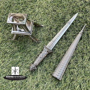 May include: A silver dagger with an ornate handle and a sheath shaped like a tower. The dagger is displayed on a silver stand shaped like the Eiffel Tower. The text "Knives Remembered - Make Your Knives Special" is visible in the bottom left corner of the image.