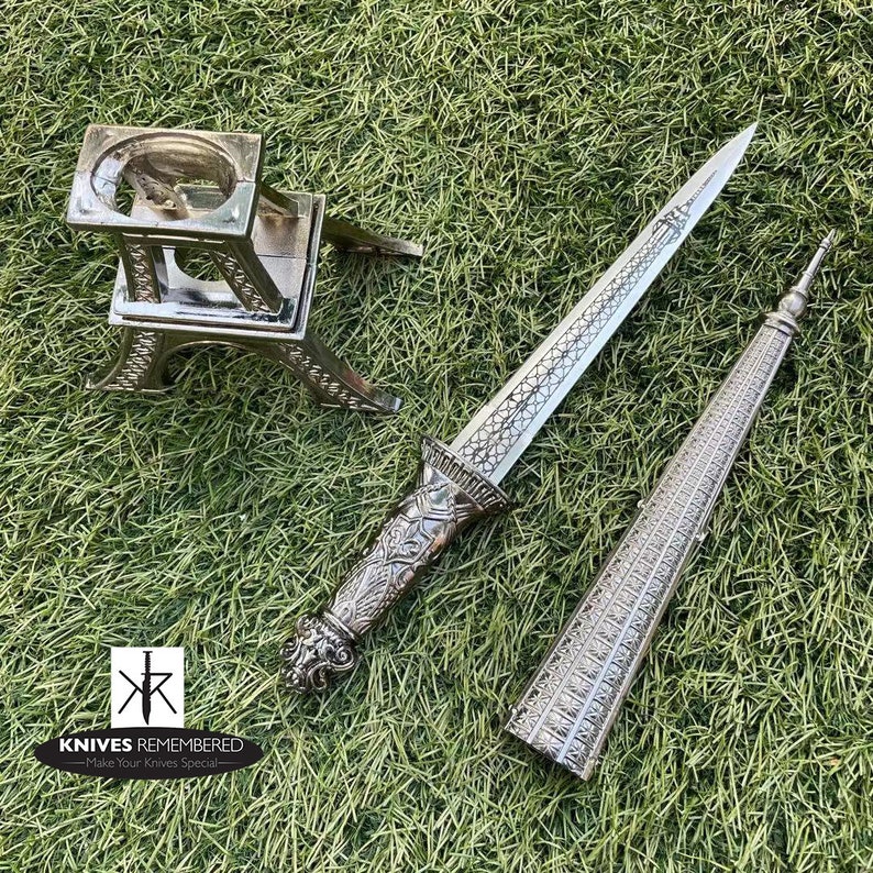 May include: A silver dagger with an ornate handle and a sheath shaped like a tower. The dagger is displayed on a silver stand shaped like the Eiffel Tower. The text "Knives Remembered - Make Your Knives Special" is visible in the bottom left corner of the image.