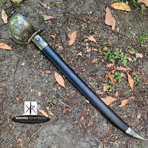 May include: A black-handled pirate sword with a gold-colored hilt featuring a ship design and a skull and crossbones. The sword is lying on a bed of brown leaves and dirt.