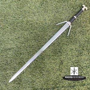 May include: A long, silver sword with a black handle and a decorative silver hilt. The sword is lying on a bed of green grass. The logo "KNIVES REMEMBERED" is in the bottom right corner.