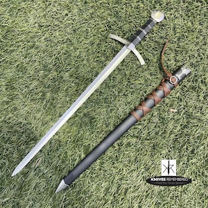 May include: A medieval-style sword with a black handle and a silver blade. The sword is lying on a green grassy surface with a brown leather scabbard next to it. The scabbard has a silver metal tip and a brown leather strap.