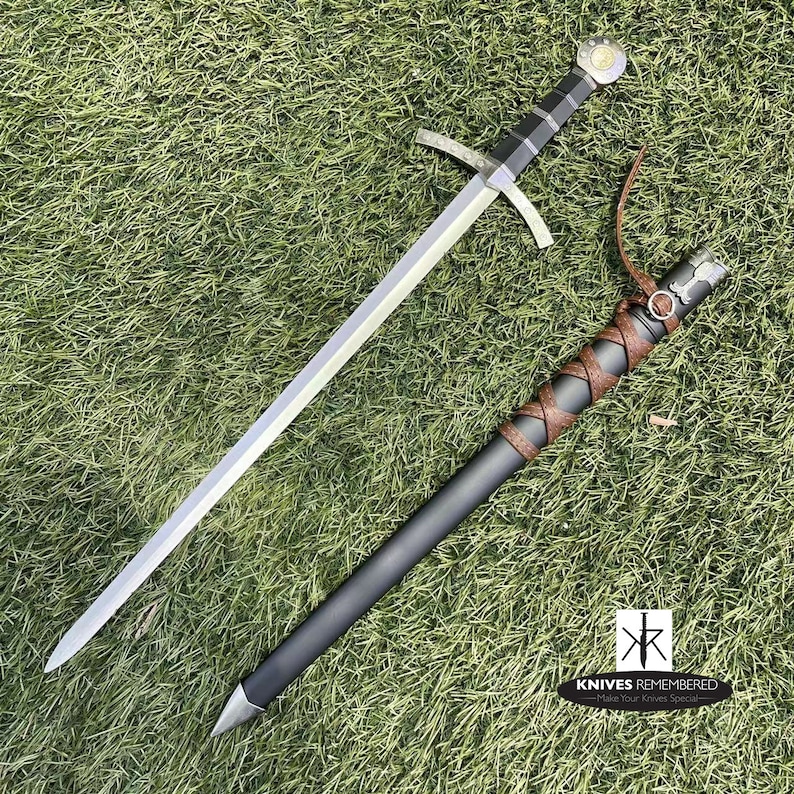 May include: A medieval-style sword with a black handle and a silver blade. The sword is lying on a green grassy surface with a brown leather scabbard next to it. The scabbard has a silver metal tip and a brown leather strap.