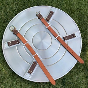 May include: A silver metal Captain America shield with brown leather straps and metal buckles. The shield is lying on a green grassy surface.